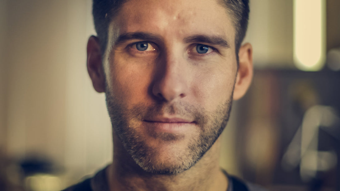 A close-up portrait of a young man with a short beard and intense gaze, hinting at a rugged handsomeness