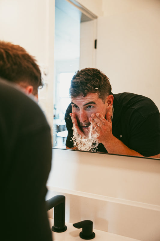 A man splashes water on his face, looking into the mirror with a focused expression, as he engages in his skincare routine.
