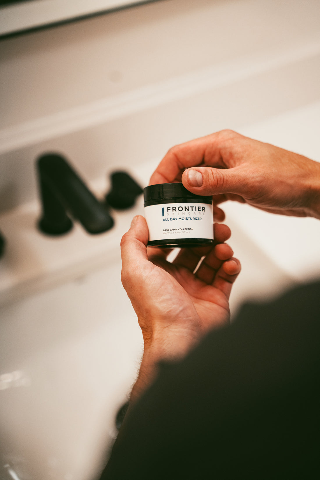  Hands hold a jar of Frontier All Day Moisturizer from their Male Care Collection, ready for use in a skincare routine.