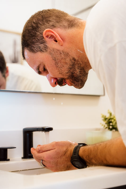 A man is washing his face over a bathroom sink, focusing intently on his skincare routine, with water droplets visible as they fall from his face. He wears a casual white t-shirt and a black wristwatch, highlighting a moment of daily grooming.