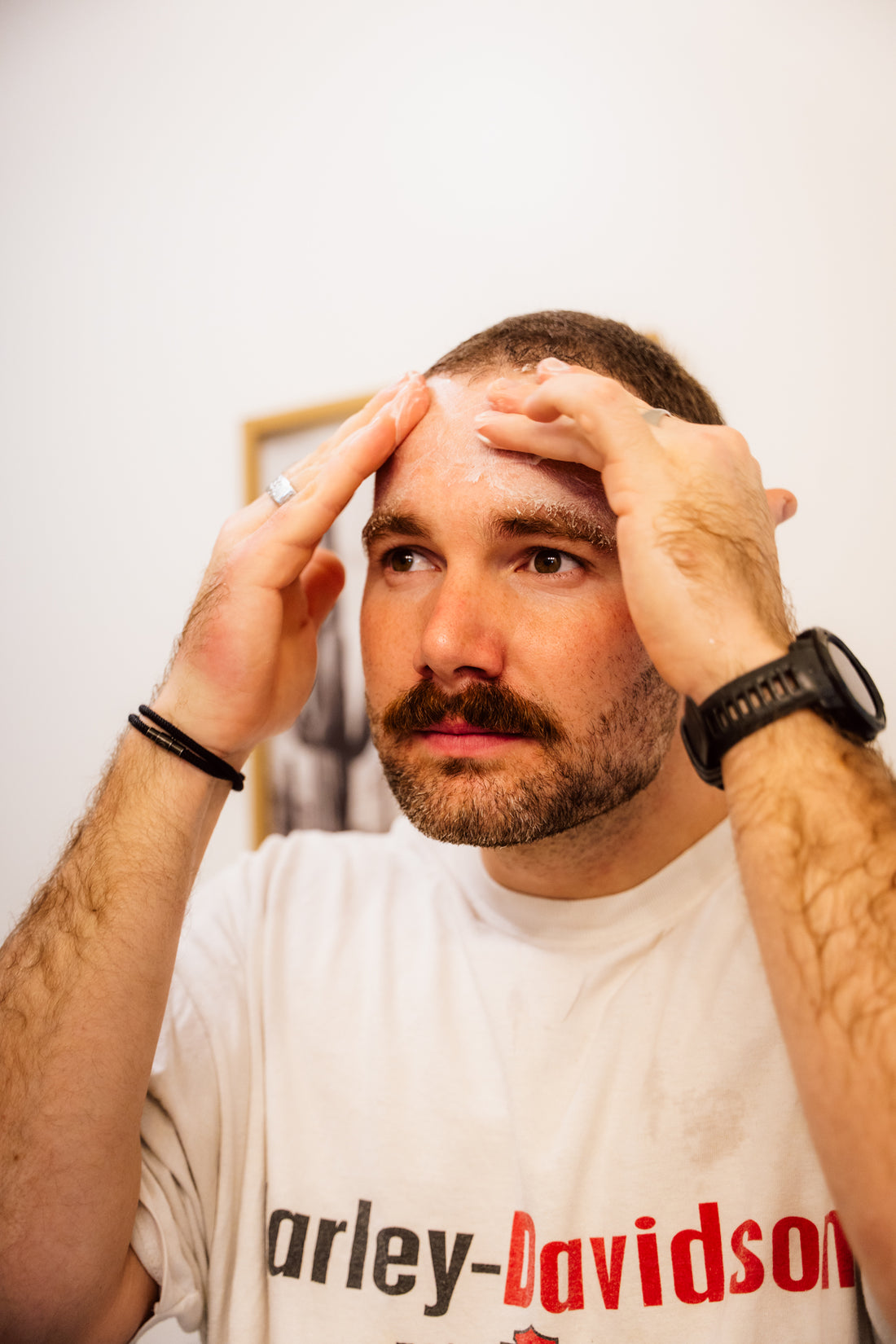 A man with a beard is examining his forehead closely, with a concerned expression possibly checking for signs of skin aging or blemishes, while wearing a Harley-Davidson t-shirt and a black wristband.