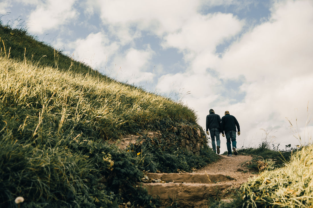 Two individuals are walking up a narrow, grassy path on a hill, their figures silhouetted against a bright cloudy sky.